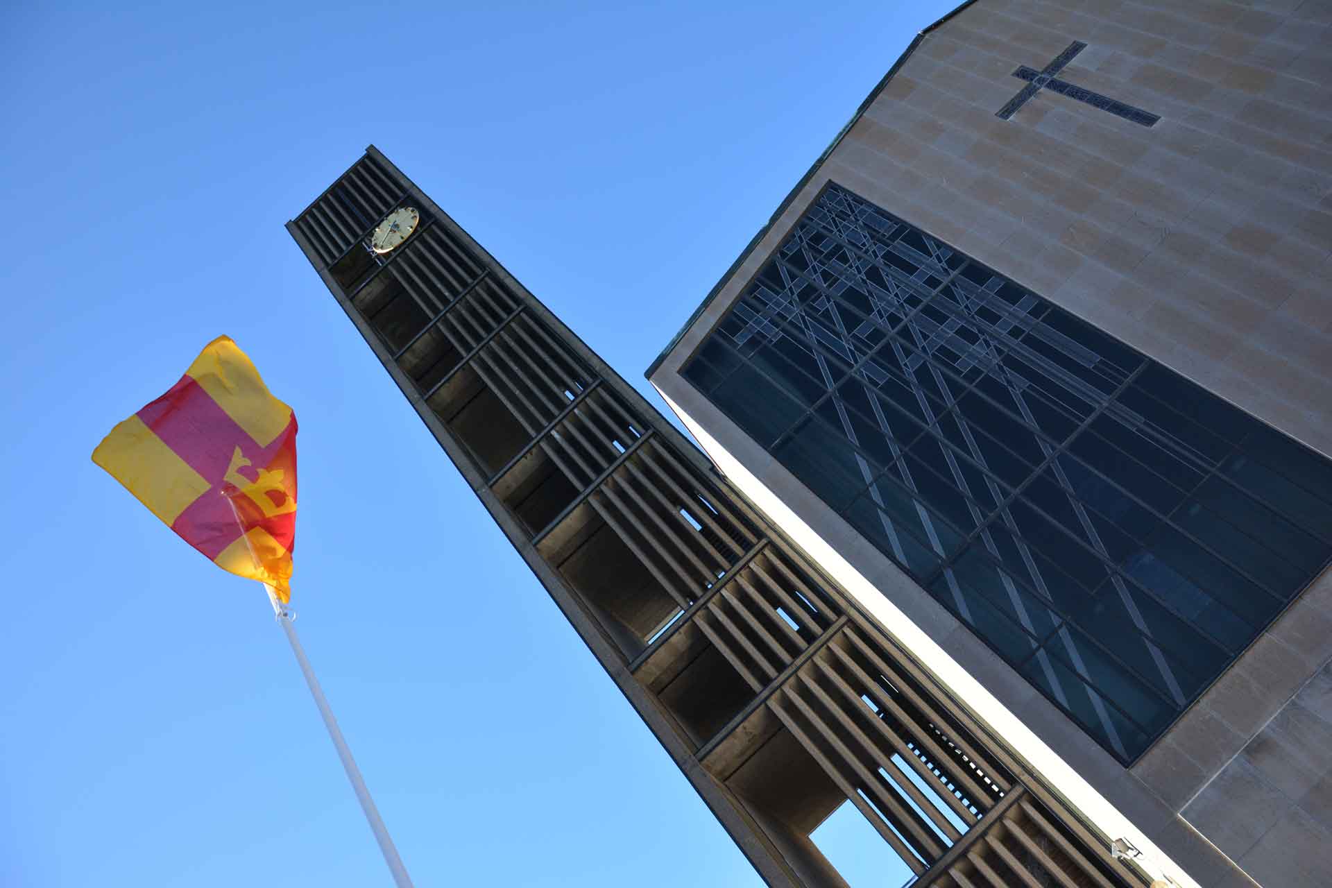 Bild på snedden där en flaggstång med rödgul flagga, klocktorn och kyrkofasad syns.