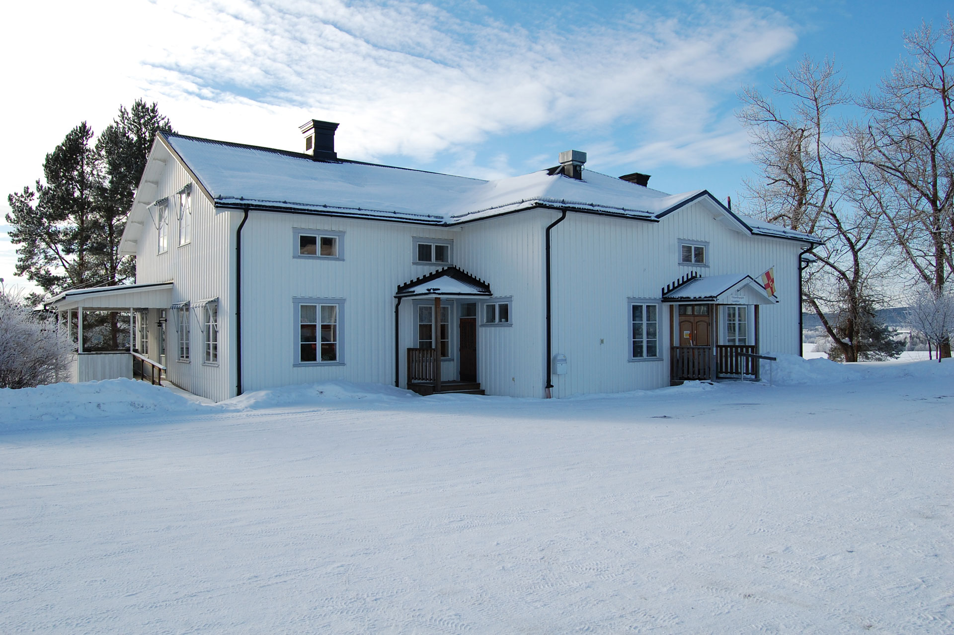 Långsele församlingshem. Vit träbyggnad. En vinterbild. Ovanför entrén hänger Svenska kyrkans flagga.
