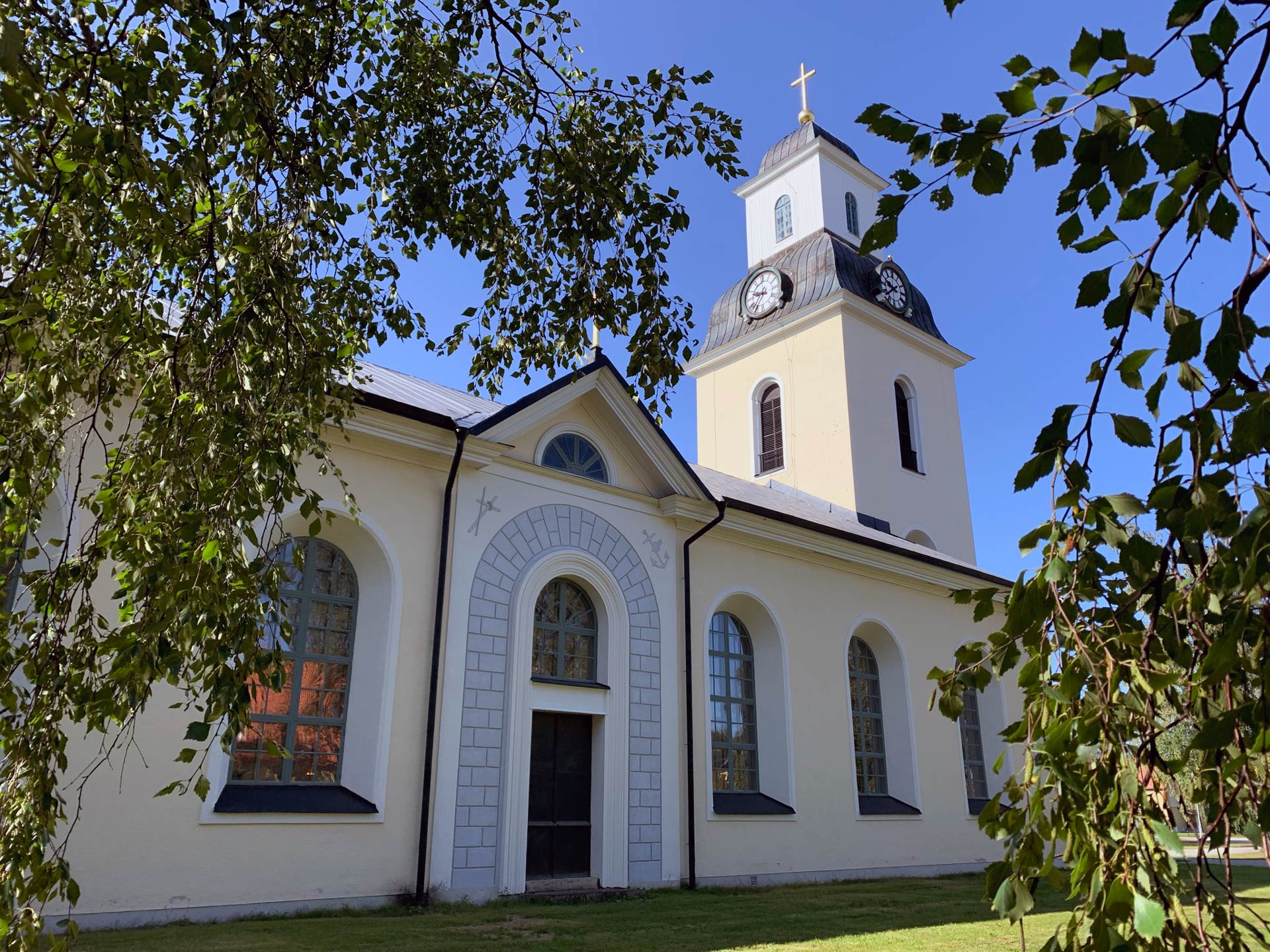 En stor stenkyrka med gulaktig puts. Det är sommar och vi ser kyrkan mellan gröna björkblad.