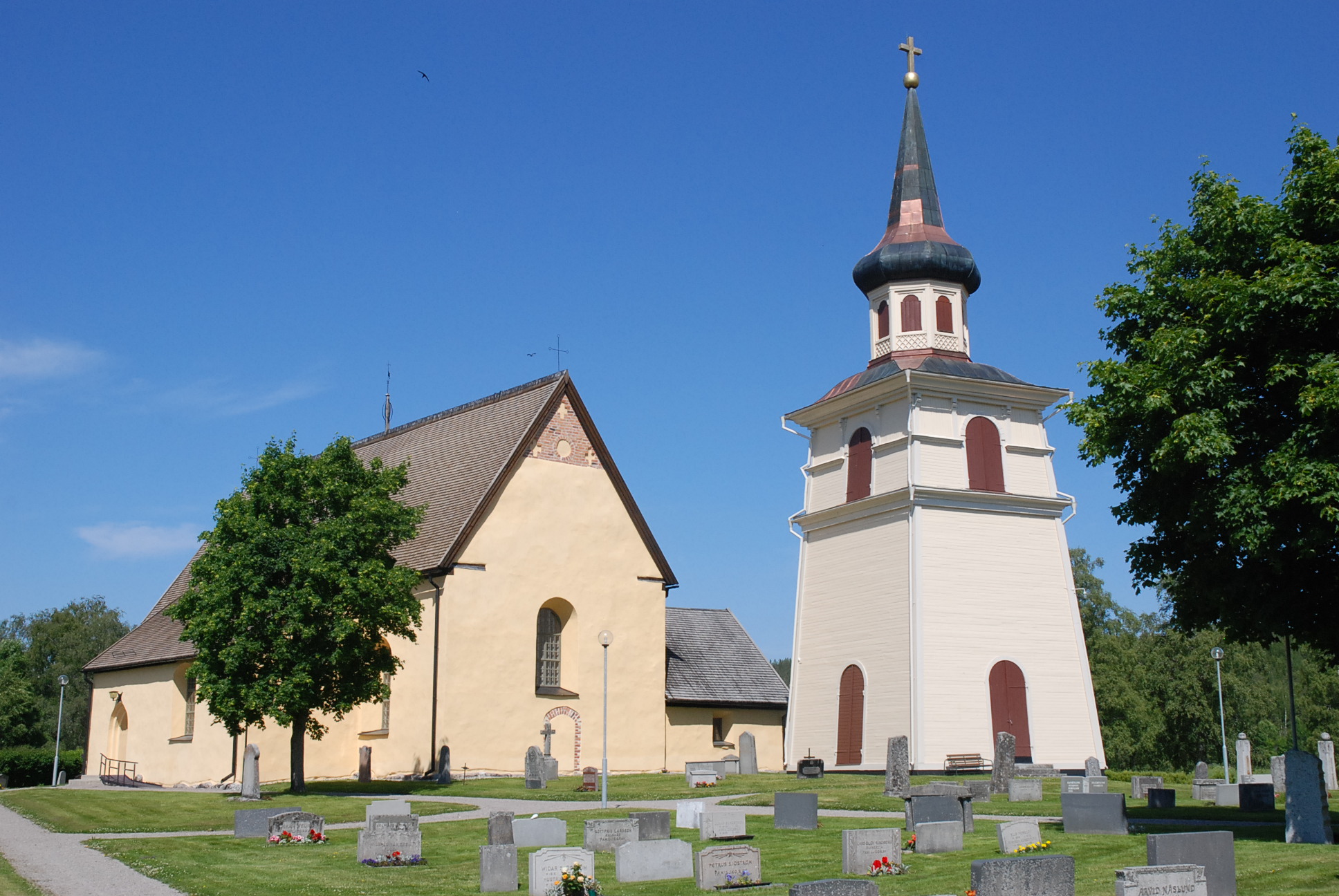 Boteå kyrka och klocktorn. I förgrunden delar av kyrkogården. Det är sommar. Mot den blå himmeln skymtar en svala.