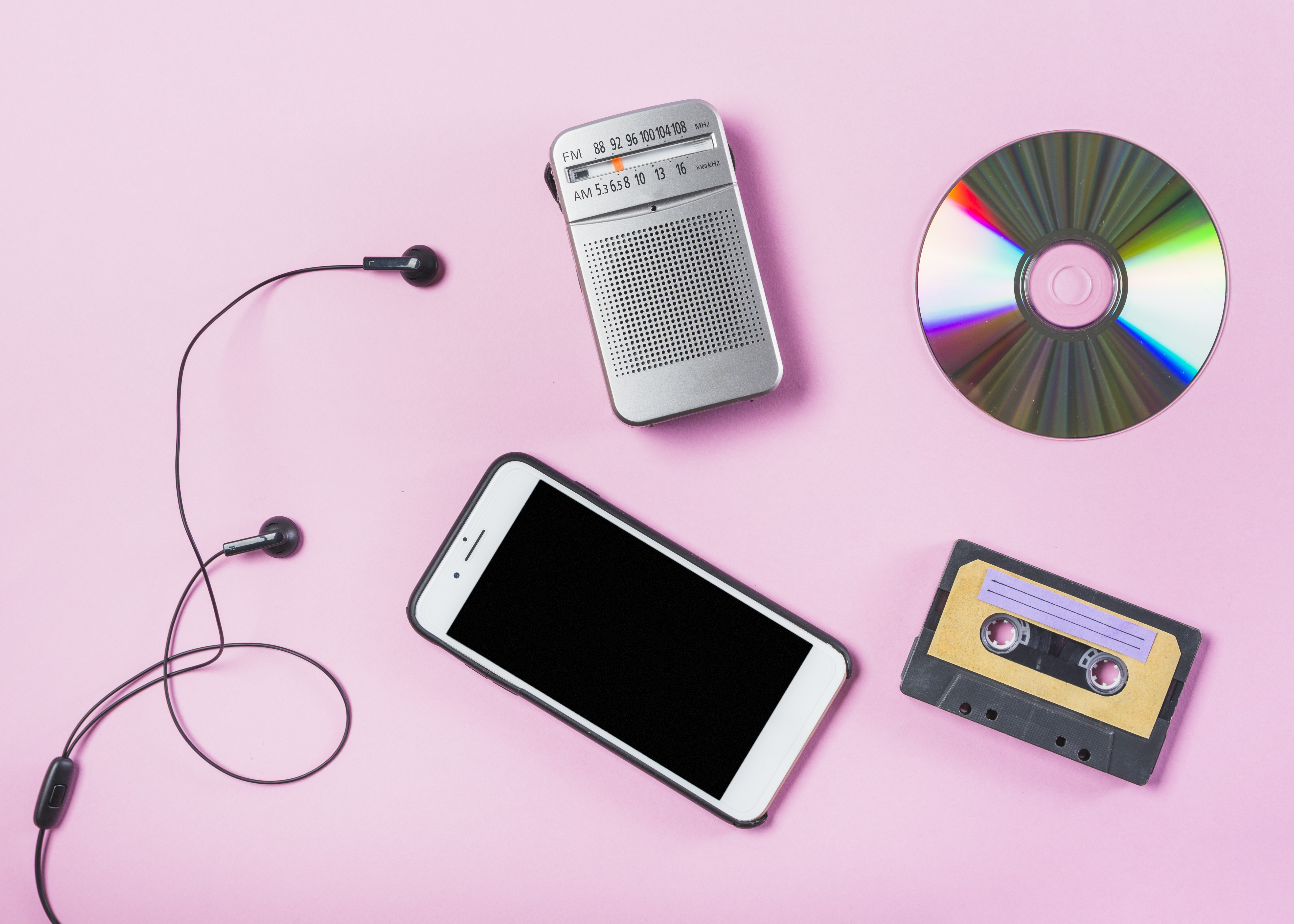 Olika musikmedier: en smartphone, ett kassettband, en cd-skiva och en fm-radio. Alla liggande på en rosa bakgrund.