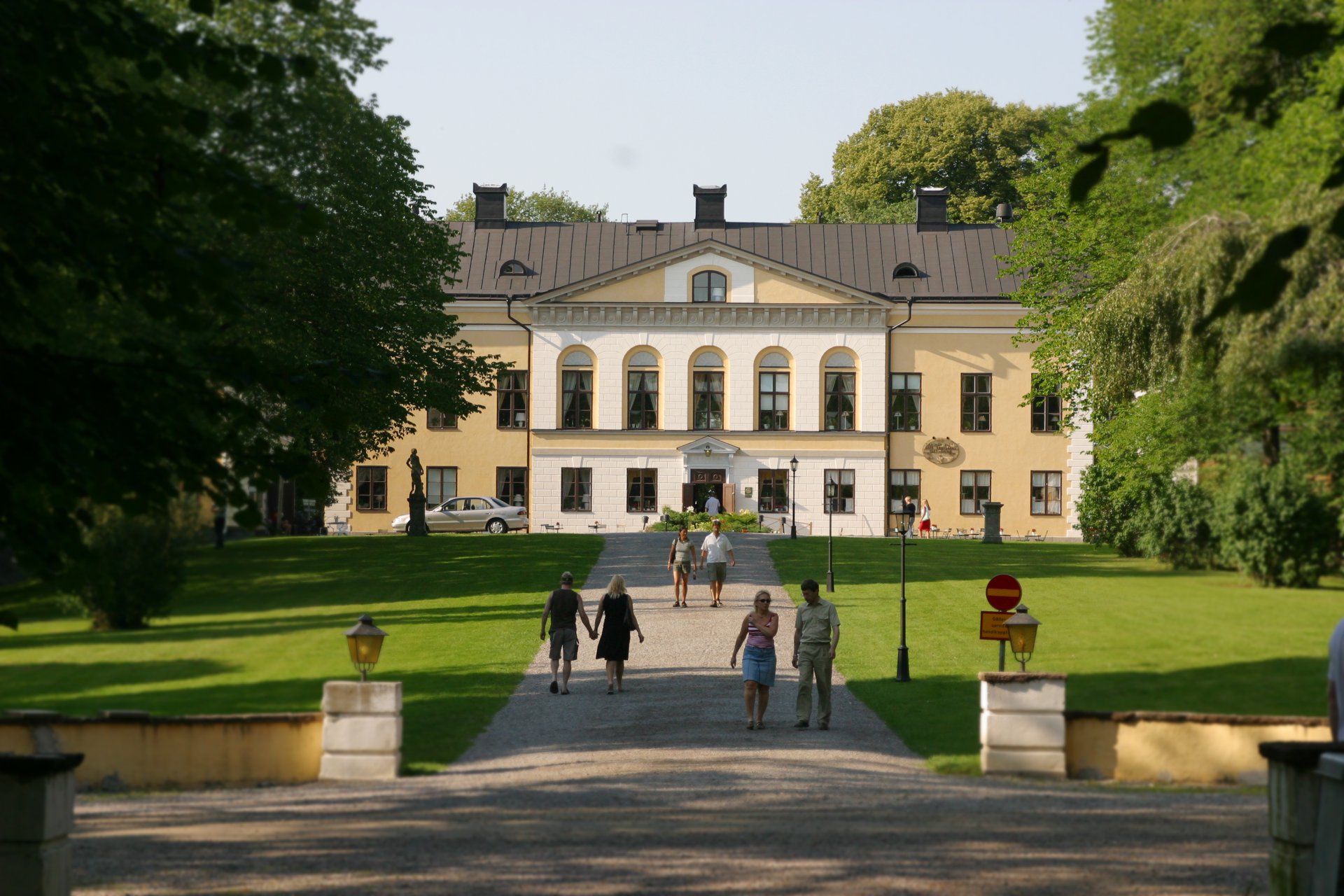 En stor beige herrgård med svart tak och många fönster. Den omges av träd. En grusgång går upp mot mitten av huset.