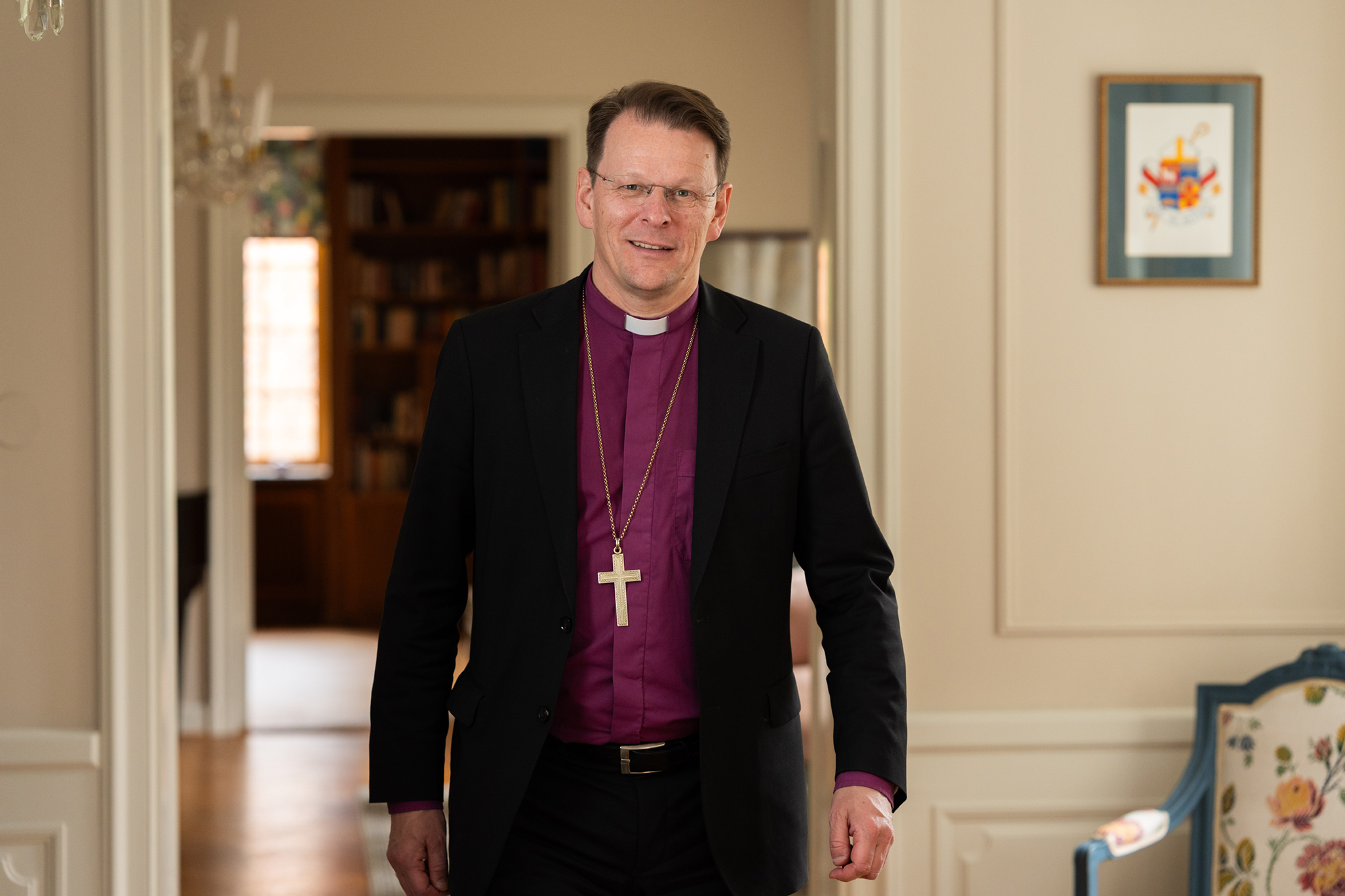 Biskop Erik står i en av salarna i Biskopgården klädd i lila  biskopsskjorta och biskopskorset kring halsen. 