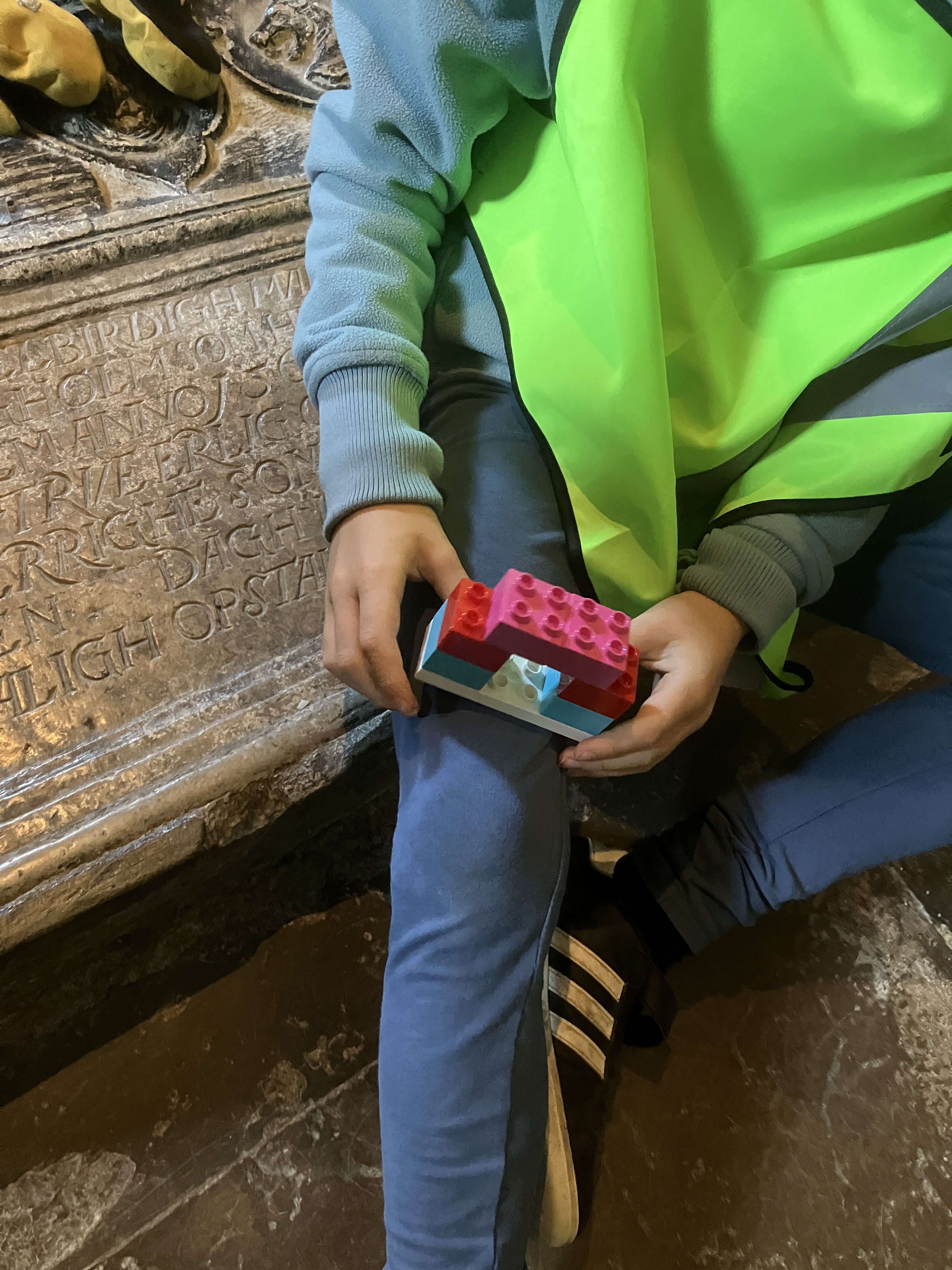 En yngre person i gul väst håller ett litet bygge av rosa, röda och ljusblå duploklossar i sitt knä.