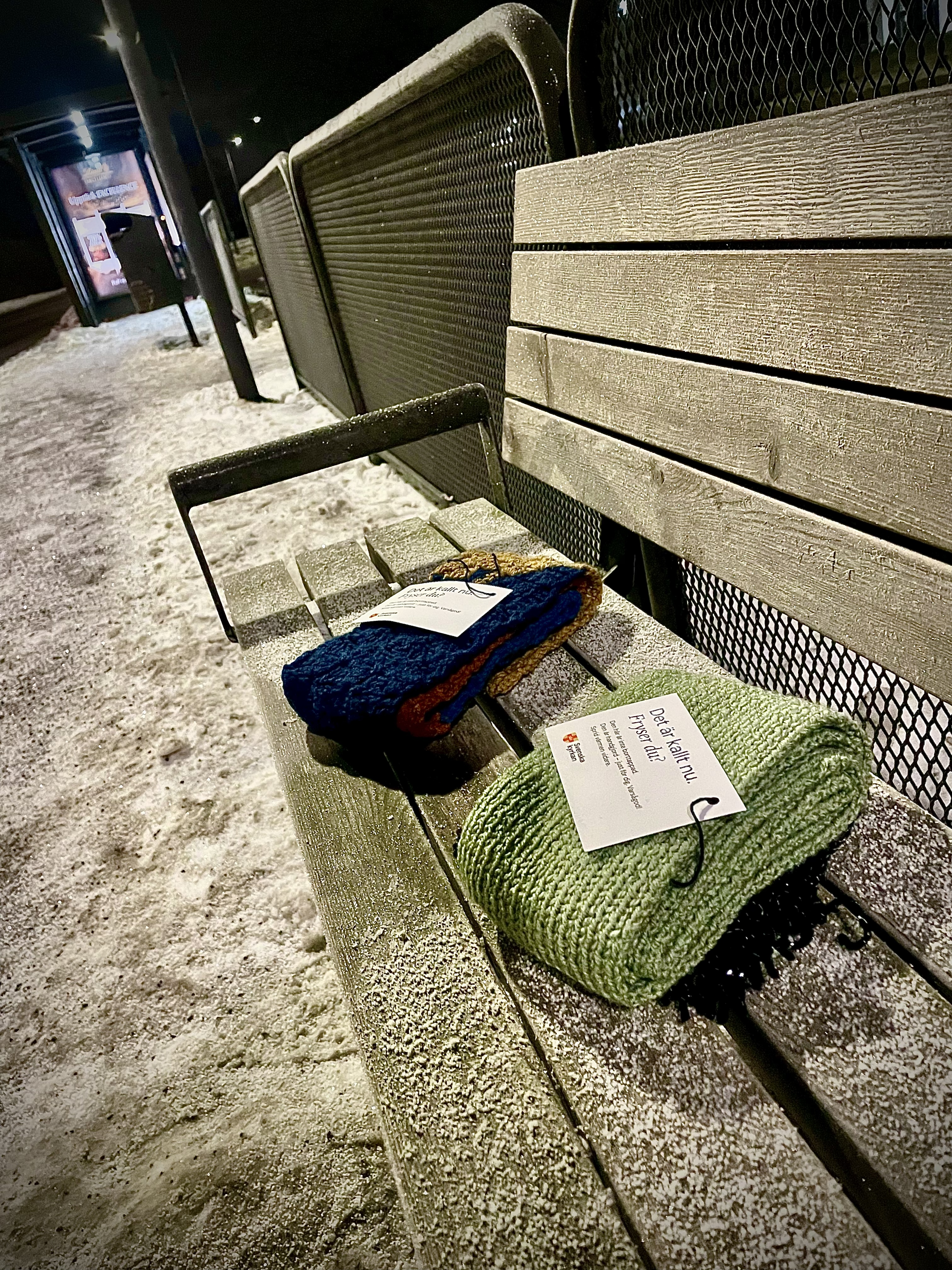 Tidig morgon. Två handgjorda halsdukar ligger på en frostig bänk vid en busshållsplats. Halsdukarna har etiketter med hälsning från Svenska kyrkan.
