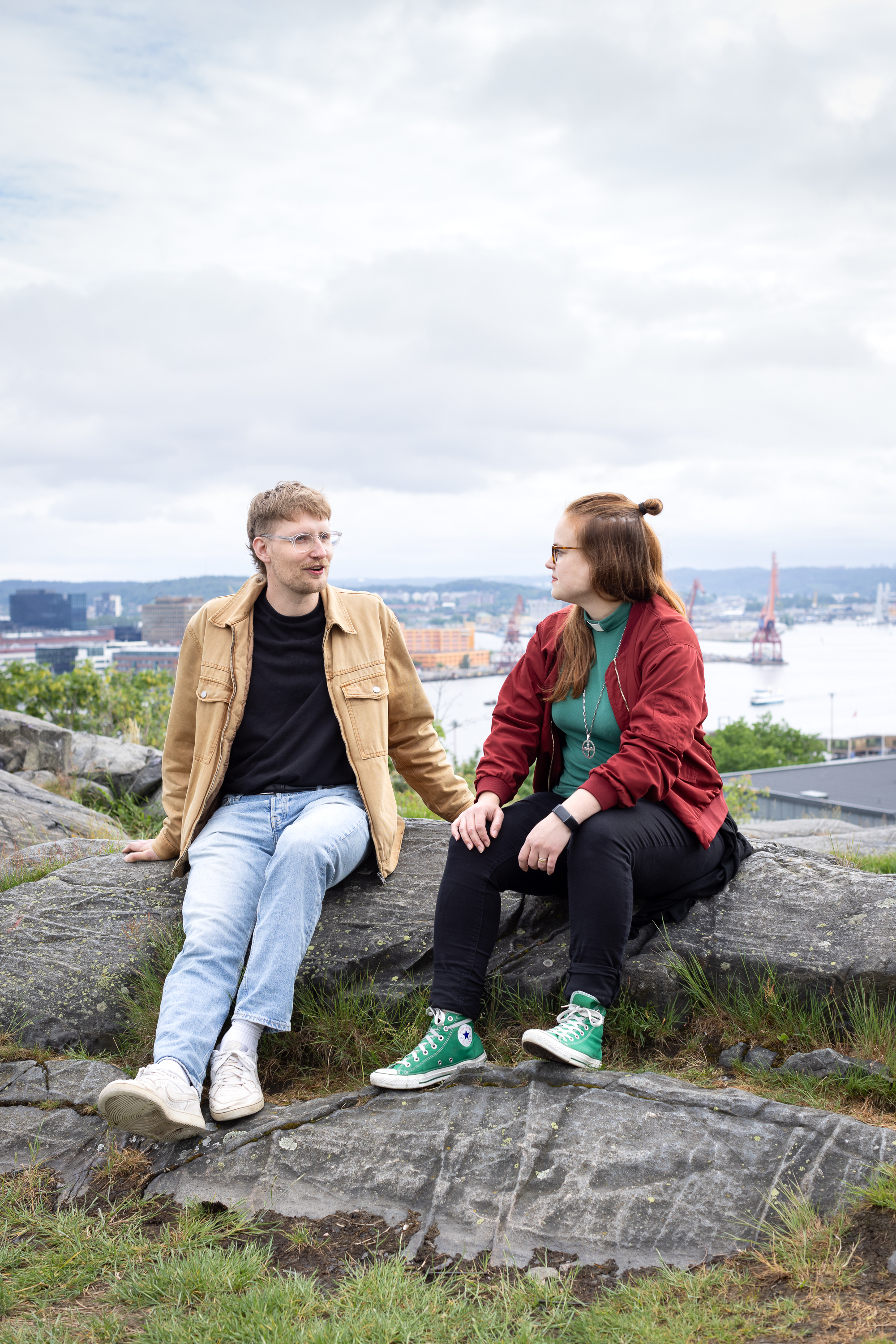 En ung man sitter på en klippa och pratar med en ung kvinna i diakonskjorta, i bakgrunden en vy över Göteborg.