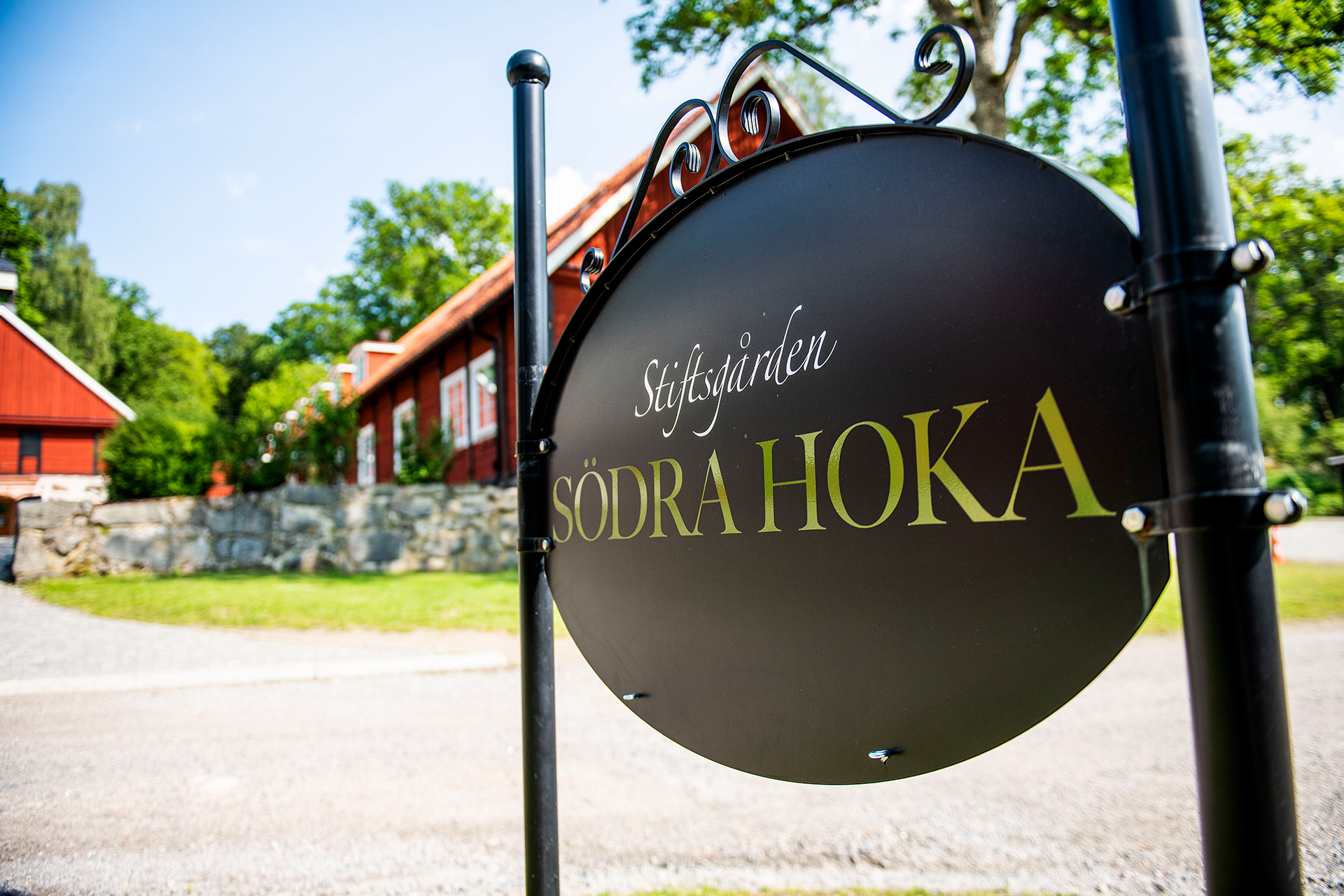 I förgrunden en svart rund skylt med texten Stiftsgården Södra Hoka. I bakgrunden syns röda trähus i lantlig miljö.