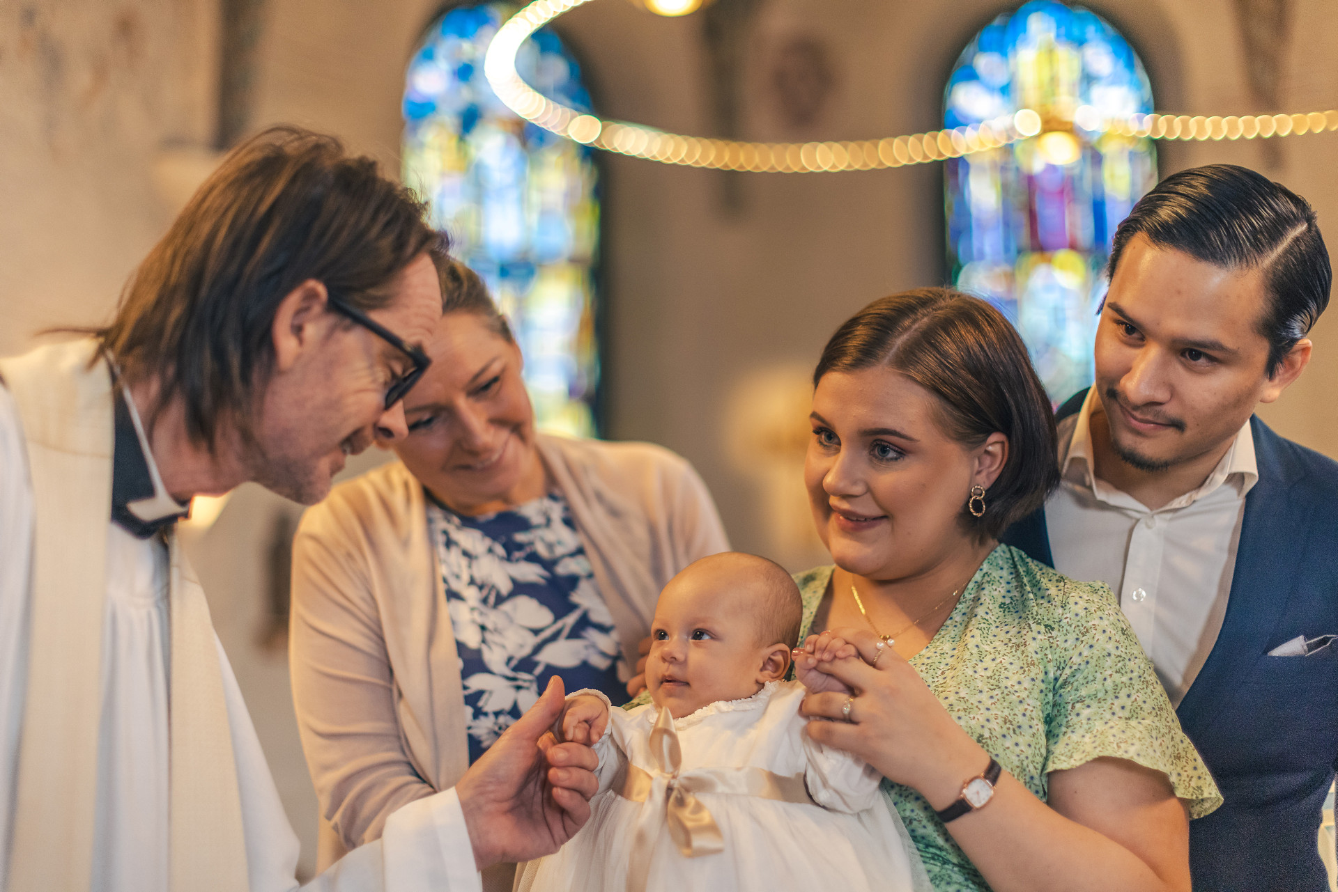 Ett par, en kvinna och en manlig präst som står med bebis i dopklädsel i kyrkan
