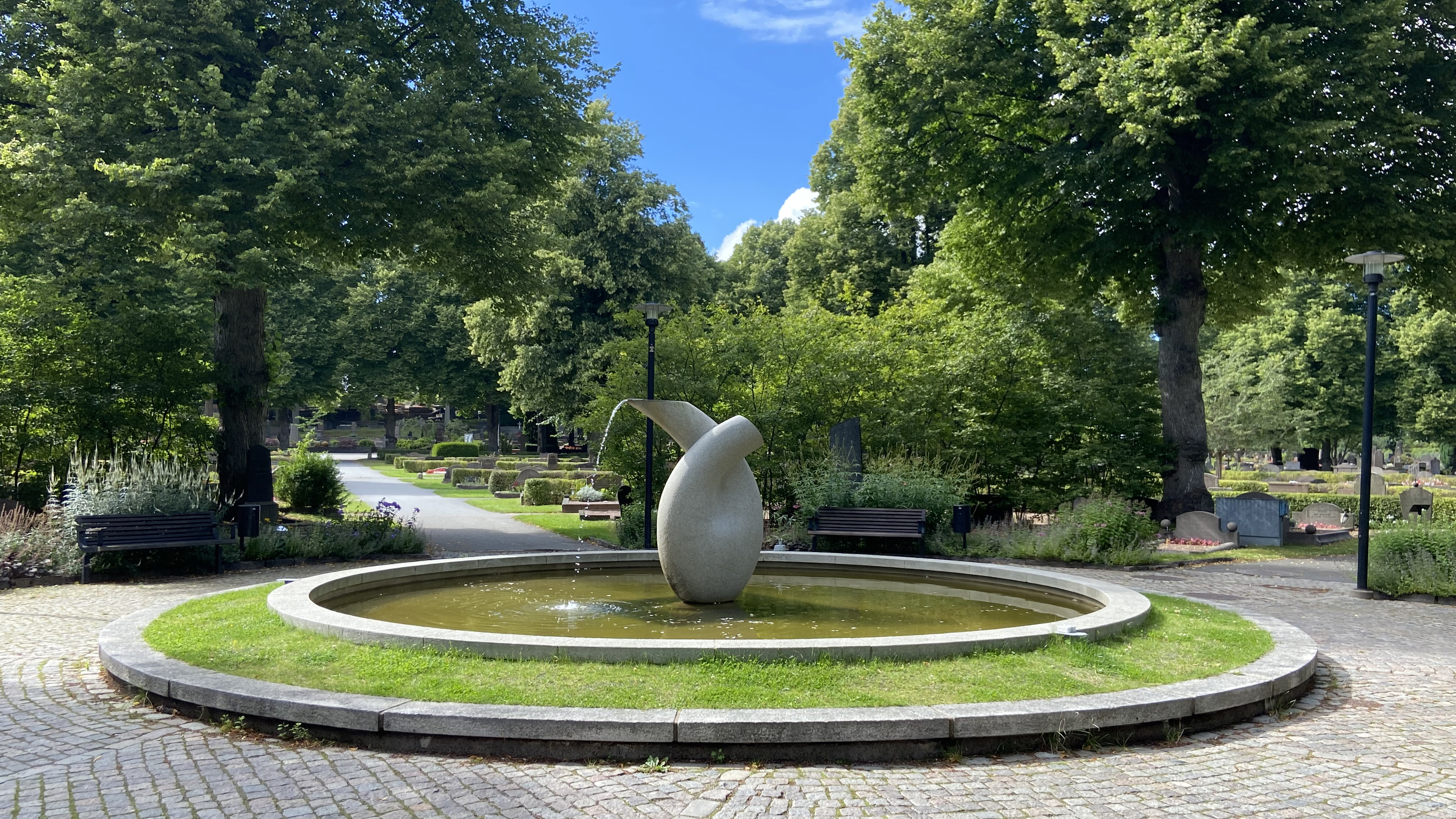 Stenskulptur med runda former som det rinner vatten ur står mitt i en stensatt damm. I bakgrunden träd, grönska och gravstenar.