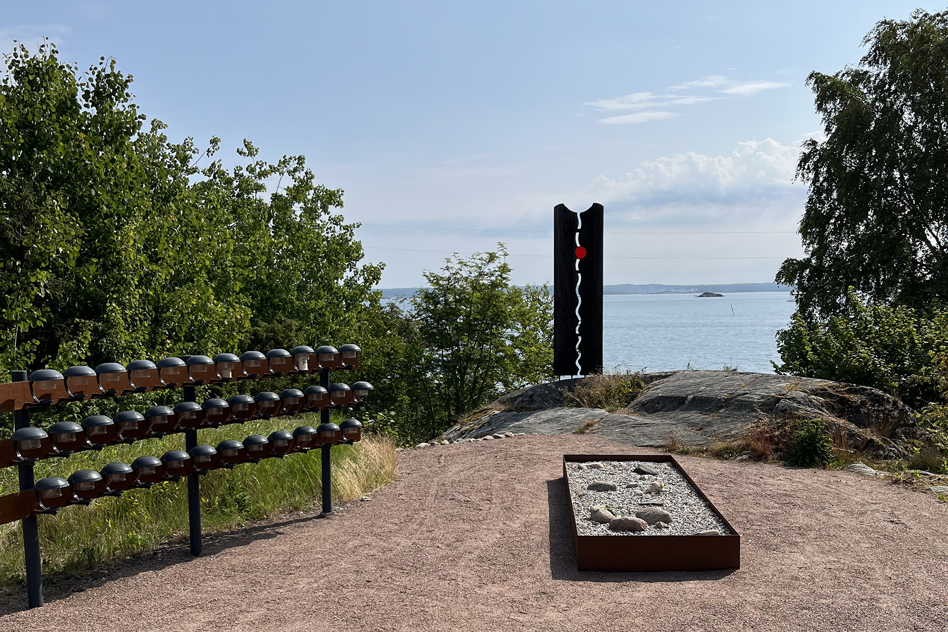 Grusad plats med havet som bakgrund. Stenstaty, ljusbärare och en corténlåda med grus och stenar.