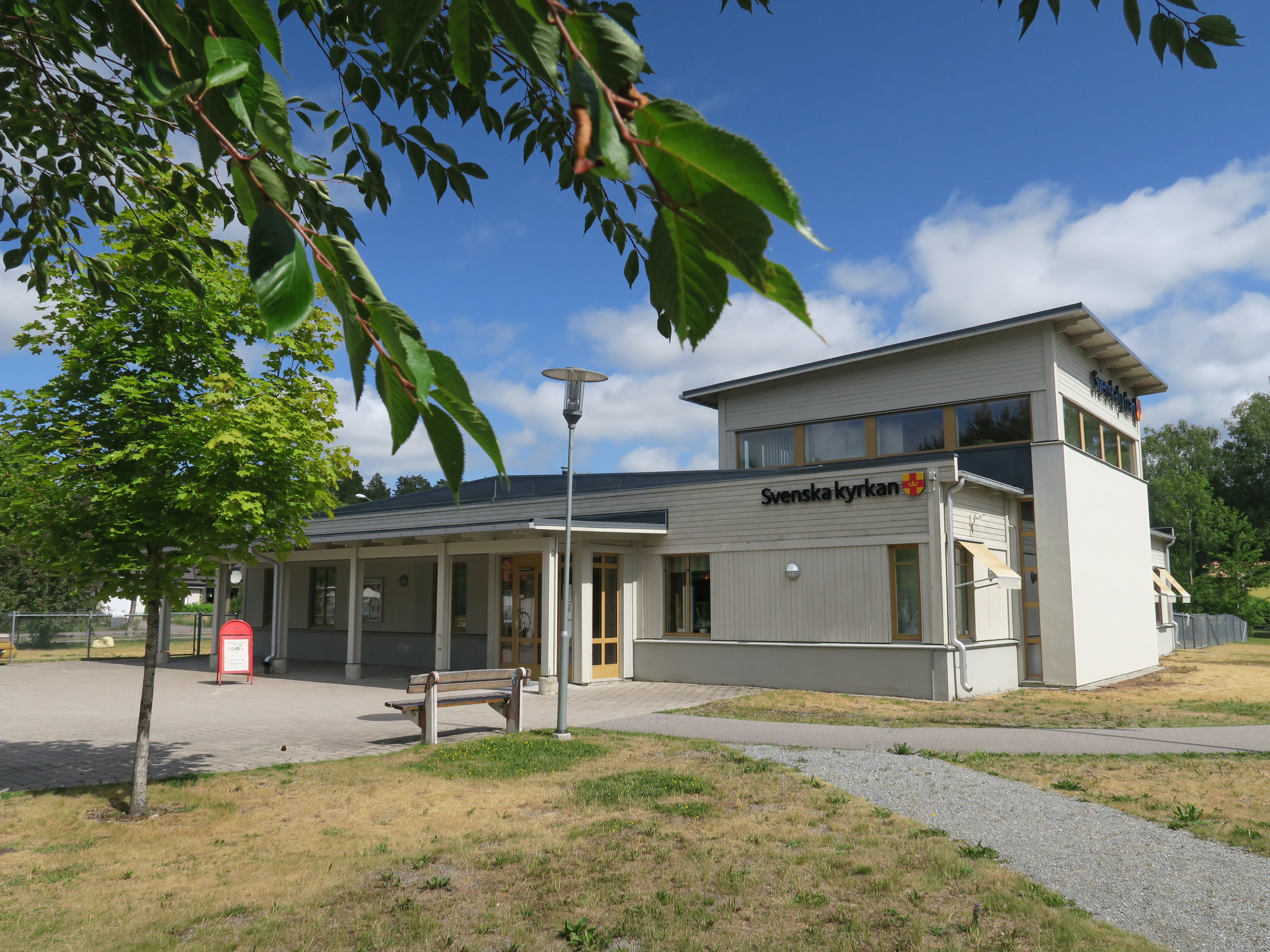 En grå byggnad med Svenska kyrkans logotyp på väggen. Runt omkring står gröna träd.