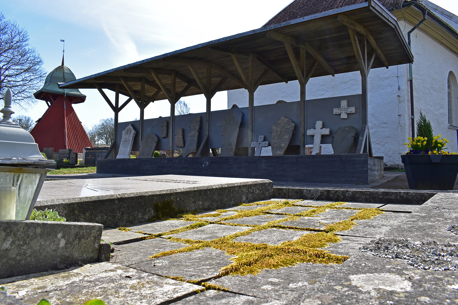 Bergums kyrkogård, lapidariets gamla gravstenar uppställda under tak  med kyrkan i bakgrunden