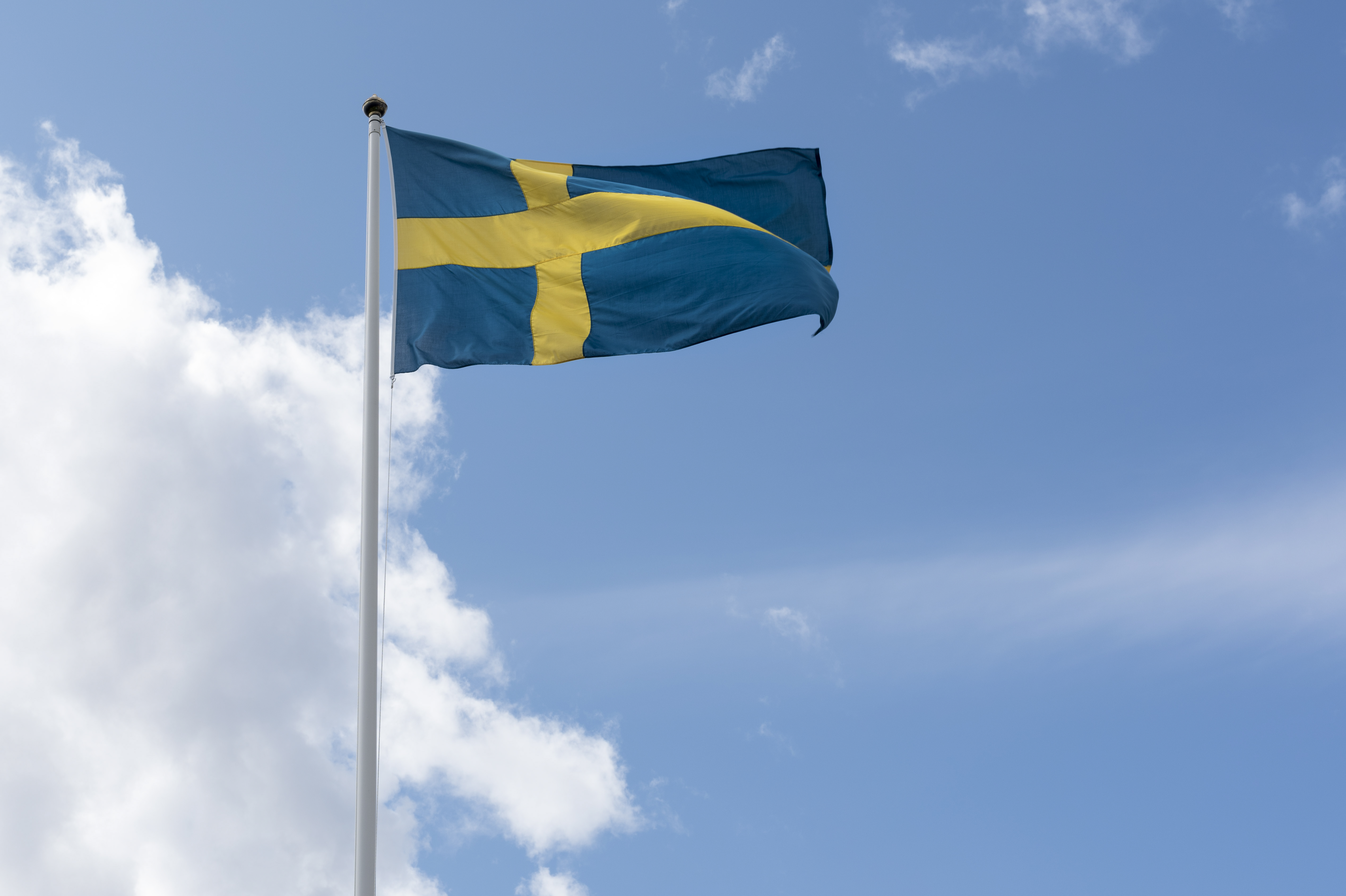En flaggstång med svenska flaggan mot blå och molnig himmel.