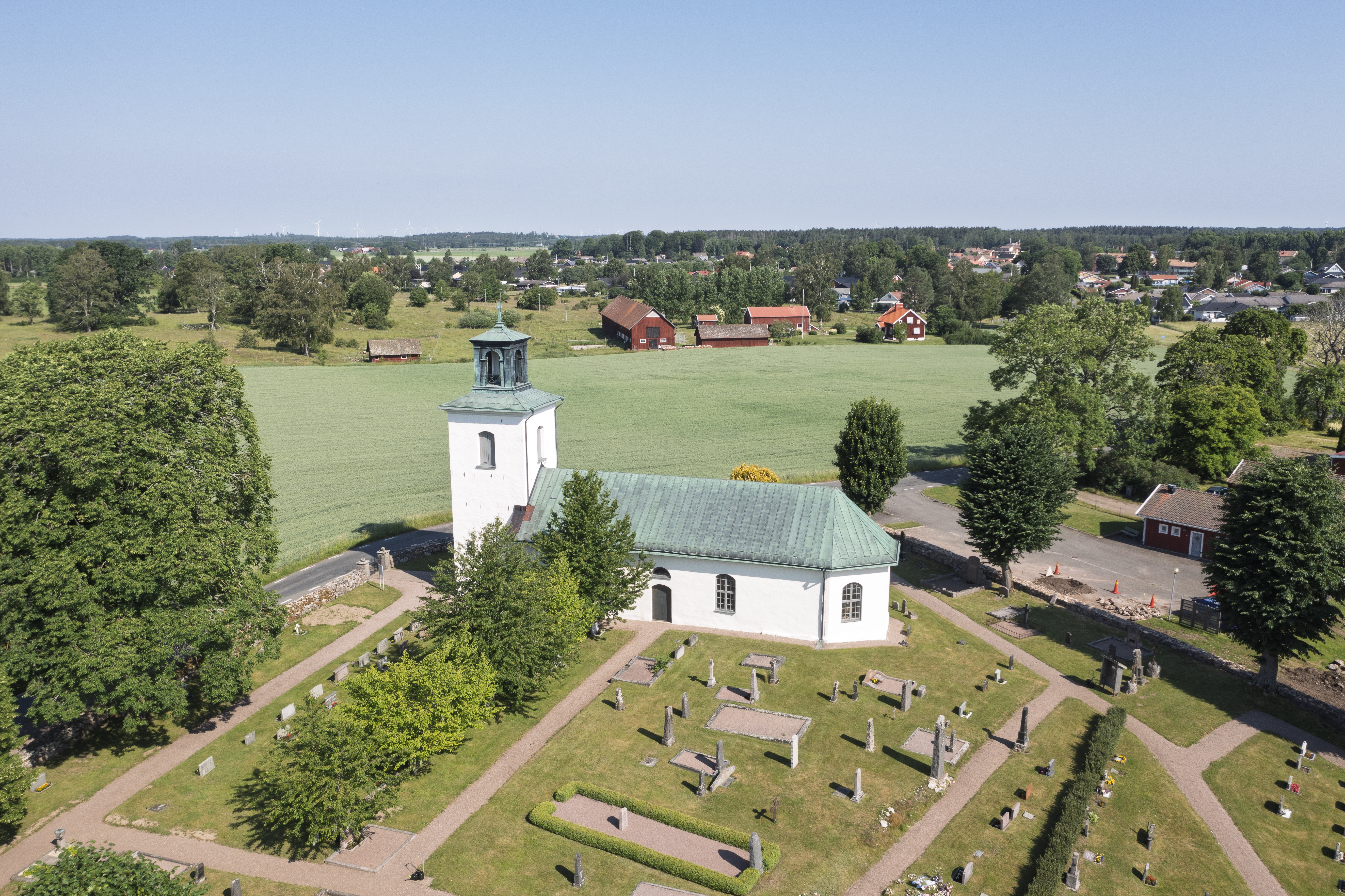 Flygfoto på en vit kyrka med grönt koppartak. Gravar, grusgångar och träd i omgivningen. En stor grön slätt i bakgrunden.