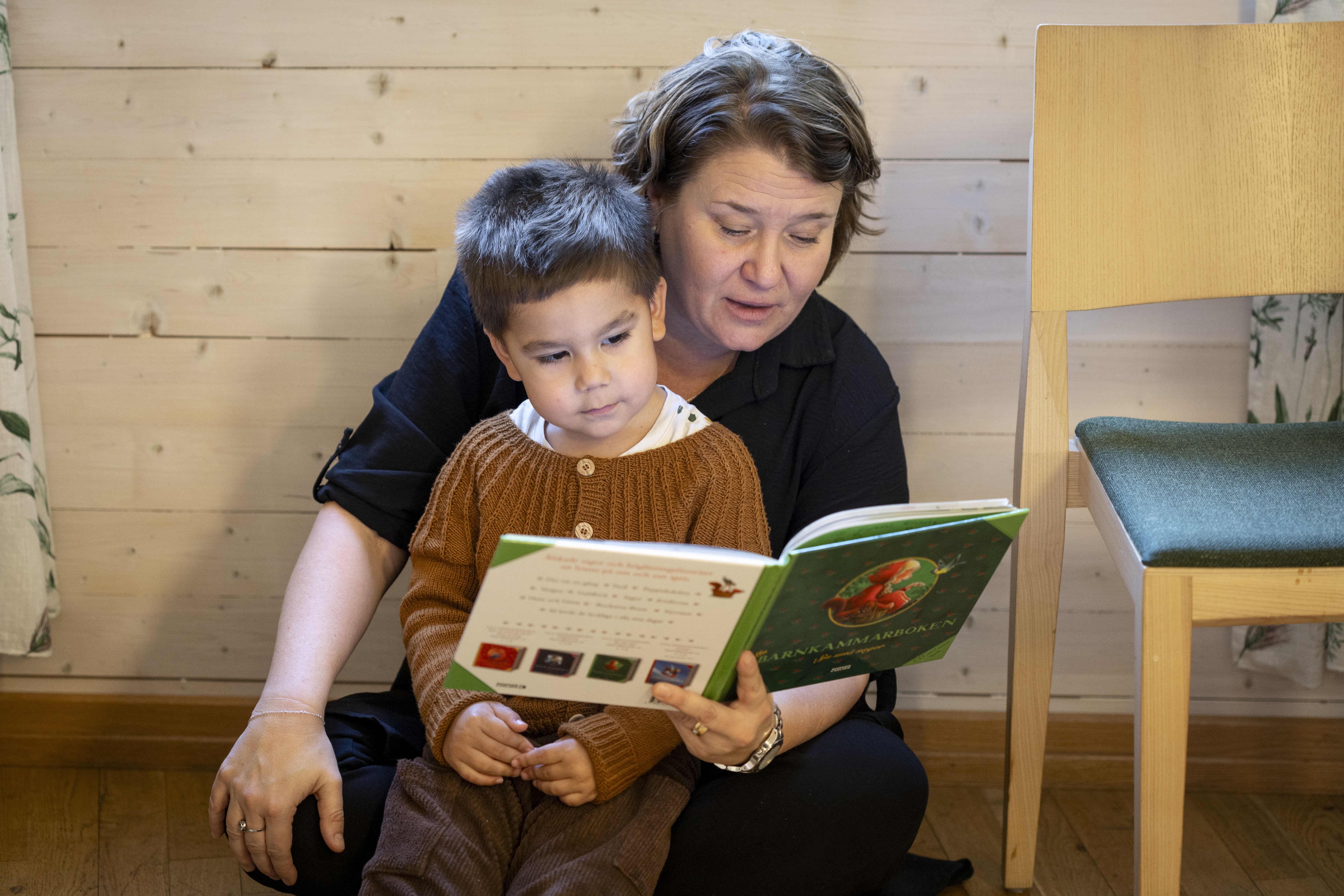 En kvinna sitter på golvet med ett barn i knät och läser en bok. En trästol med grön sittdyna står bredvid.