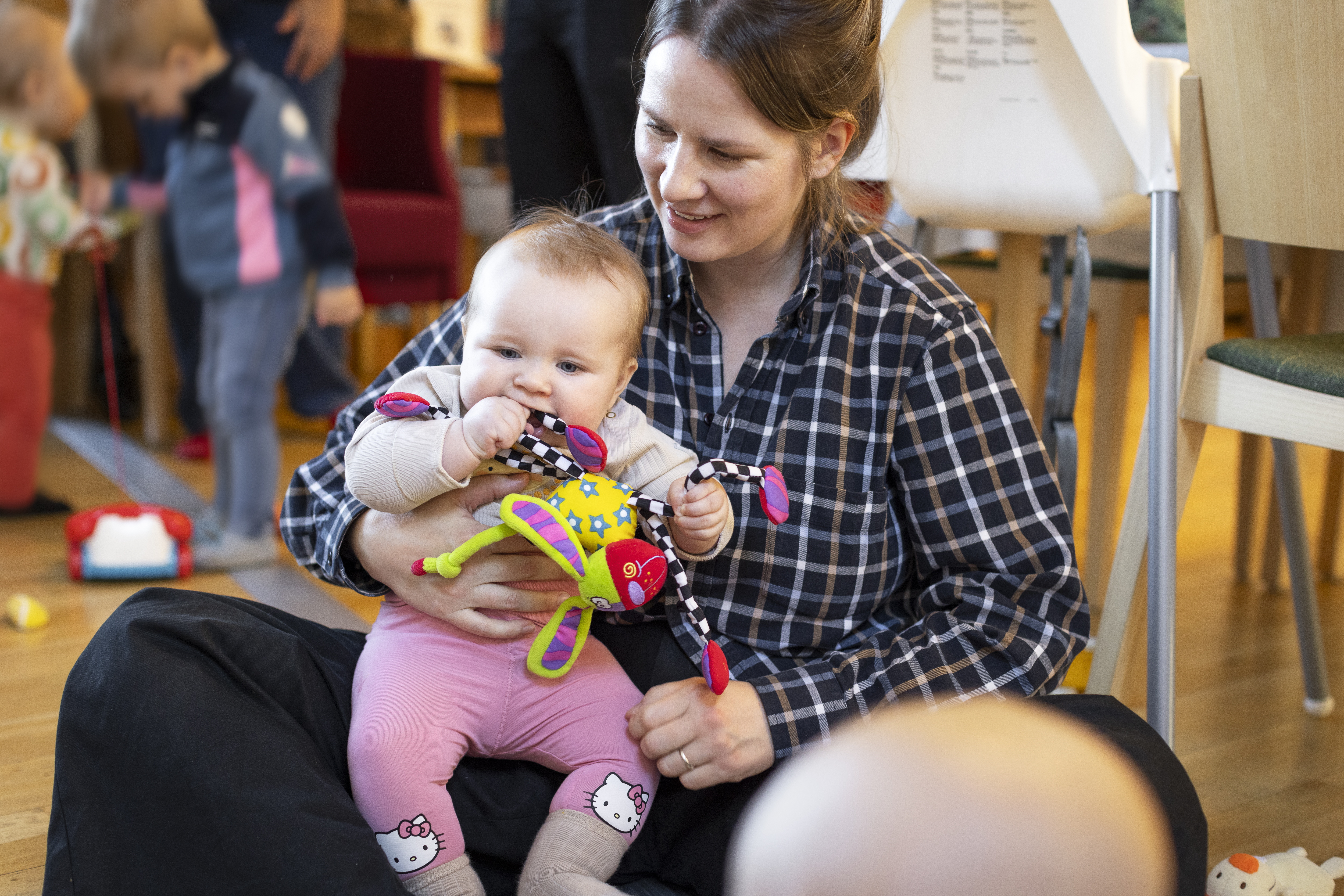 En kvinna i rutig skjorta sitter på golvet med en bebis i famnen. Bebisen tuggar på ett mjukt gosedjur.