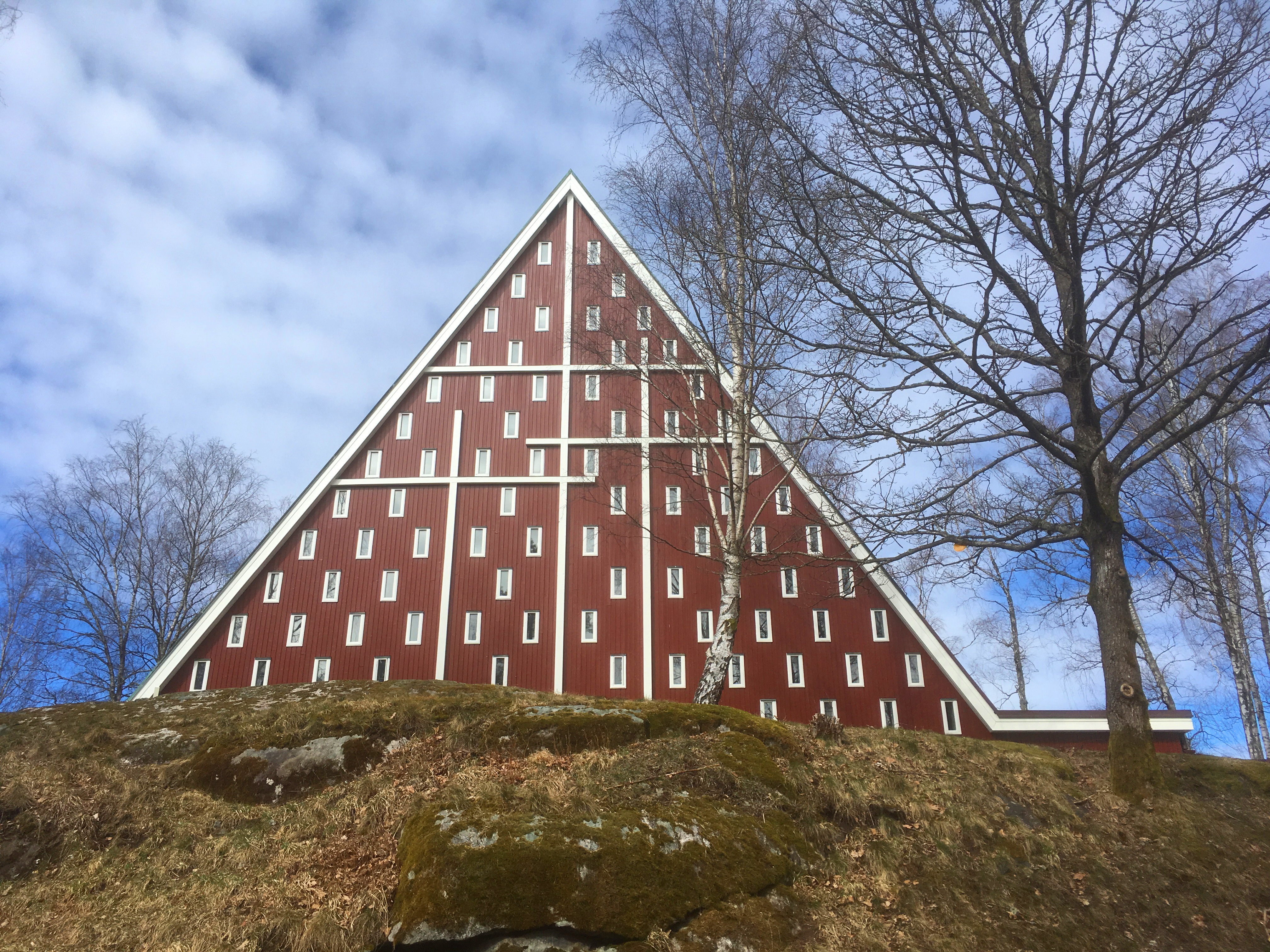 Röd träkyrka med tre vita kors på gaveln. Omgiven av träd utan löv.